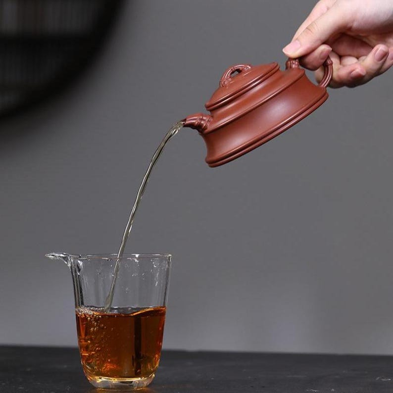 May include: A dark reddish-brown clay teapot with a flat lid and a curved handle pouring amber-colored tea into a clear glass pitcher. The teapot has a smooth, matte finish and a distinct tiered design on the body, while the glass pitcher is transparent.
