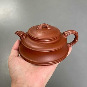May include: A reddish-brown clay teapot featuring a wide, flat base and a tiered body design. The lid is adorned with a sculpted bamboo leaf motif, while the spout and handle are shaped to resemble segmented bamboo stalks with a smooth finish.