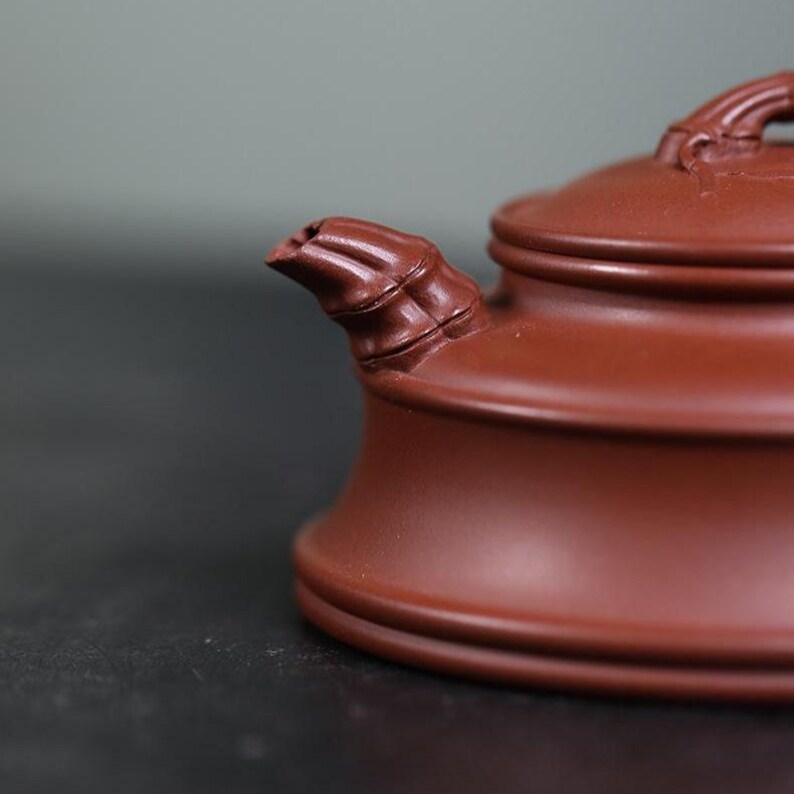 May include: A reddish-brown clay teapot featuring a spout and lid handle sculpted to resemble bamboo segments. The teapot has a smooth, matte finish with a wide, circular base and a tiered lid design, showcasing detailed craftsmanship.
