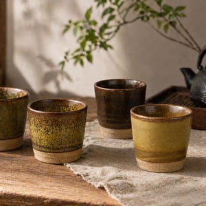 以下が含まれることがあります： 緑と茶色の手作りセラミックティーカップ4個。各カップは、テクスチャ加工された仕上げで、明るい色のベースと素朴でアースカラーの美しさがあります。カップは、ニュートラルカラーの布が敷かれた木製の表面に配置されています。