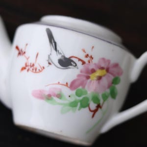 May include: White ceramic teapot with a painted design of a bird and flowers. The bird is black and gray, and the flowers are pink, yellow, and green. The teapot has a handle and a spout.