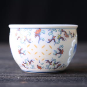 Może przedstawiać: Mała, biała ceramiczna miska z niebieską obw&oacute;dką i skomplikowanymi wzorami kwiatowymi w kolorze pomarańczowym, niebieskim i ż&oacute;łtym. Miska ma zaokrąglony kształt i dekoracyjny design, idealna do serwowania małych porcji.