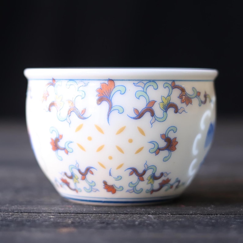 Może przedstawiać: Mała, biała ceramiczna miska z niebieską obw&oacute;dką i skomplikowanymi wzorami kwiatowymi w kolorze pomarańczowym, niebieskim i ż&oacute;łtym. Miska ma zaokrąglony kształt i dekoracyjny design, idealna do serwowania małych porcji.