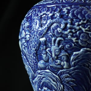 Könnte beinhalten: Eine tiefblaue und weiße Keramikvase mit komplexen Reliefschnitzereien einer ziegenartigen Figur, stilisierten Bäumen und menschenähnlichen Charakteren. Die Oberfläche weist ein glänzendes Finish mit detaillierten Texturen auf.