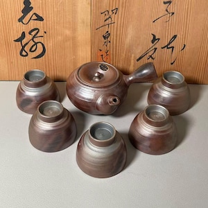 Può includere: Un servizio da tè in ceramica marrone con una teiera e sei tazze abbinate. La teiera ha un manico e un coperchio. Le tazze sono piccole e rotonde. Il set è esposto su una superficie grigio chiaro.