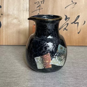Op de afbeelding: Een zwarte keramische sake-kan met een getextureerd oppervlak en een gouden rand. De kan heeft decoratieve metalen vierkanten in goud, roze en groen. De achtergrond bevat houten panelen met Japanse kalligrafie.