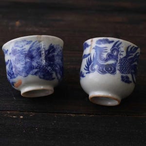 Puede incluir: Dos pequeñas tazas de cerámica blanca con diseños de dragones azules. Las tazas tienen forma redondeada y un borde ligeramente ensanchado. El dragón está representado en un estilo chino tradicional. Las tazas son probablemente para té o sake.