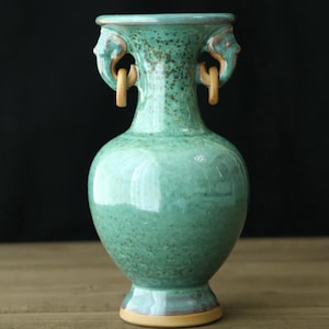 Op de afbeelding: Een turquoise keramische vaas met een gespikkelde afwerking. De vaas heeft twee decoratieve handvatten met een gezichtsontwerp en een lichtbruine ring. De basis van de vaas heeft een lichtbruine rand. De achtergrond is zwart.