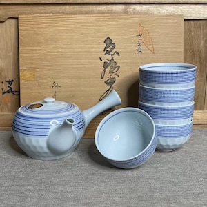 May include: A Japanese tea set with a blue and white striped design. The set includes a teapot, a stack of four small bowls, and one additional bowl. The set is displayed in front of a wooden box with Japanese characters.