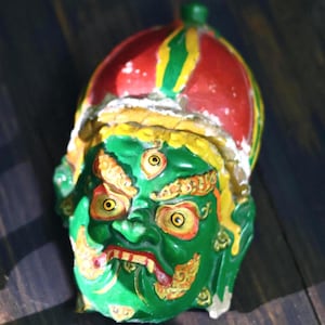 May include: A green, hand-painted mask with a fierce expression. The mask features a red and yellow headdress, yellow accents, and orange and yellow details around the eyes and mouth. The mask is a unique decorative item.