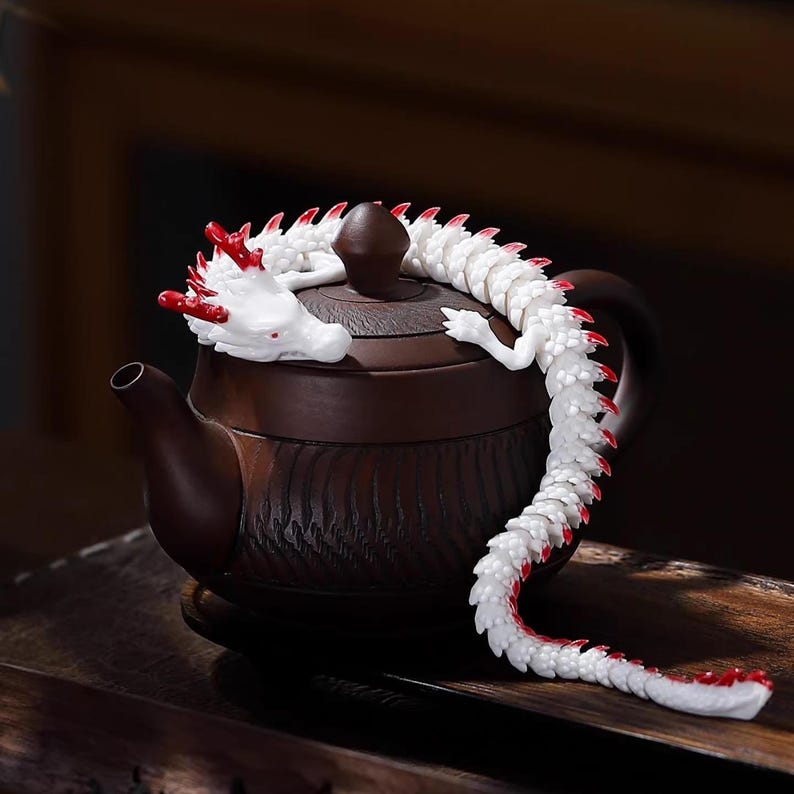 May include: A brown ceramic teapot with a decorative white and red dragon wrapped around the lid. The dragon's head is white with red accents, and its body is white with red spines. The teapot sits on a wooden surface.