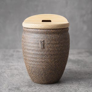 May include: A brown ceramic jar with a bamboo lid. The jar has a textured surface and a slightly rounded shape. The lid has a rectangular opening. The jar appears to be handmade and has a rustic aesthetic.
