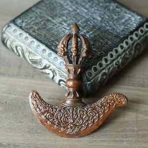 May include: A brown, ornate ritual knife with a crown-like handle. The curved blade features detailed scrollwork. The object is set against a decorative box on a wooden surface, suggesting a spiritual or ceremonial theme.