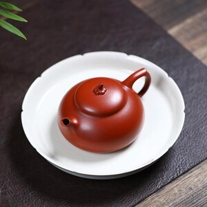 May include: A small, round, reddish-brown teapot with a matching lid and handle, resting on a white, scalloped-edge plate. The teapot has a small spout and a decorative knob on the lid. The plate sits on a dark surface.