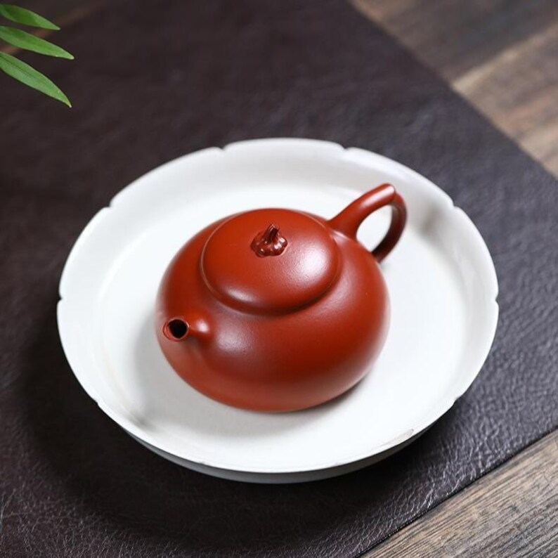 May include: A small, round, reddish-brown teapot with a matching lid and handle, resting on a white, scalloped-edge plate. The teapot has a small spout and a decorative knob on the lid. The plate sits on a dark surface.