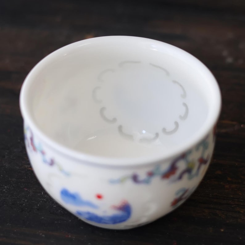 Może przedstawiać: Mała, biała ceramiczna miska z kwiatowym wzorem w kolorze niebieskim, czerwonym i zielonym. Wnętrze ma słaby, okrągły wz&oacute;r. Misa stoi na ciemnej drewnianej powierzchni.