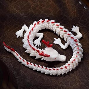 May include: A white dragon figurine with red accents. The dragon is curled up, with its head and tail close together. The figurine has detailed scales and is posed on a brown surface.