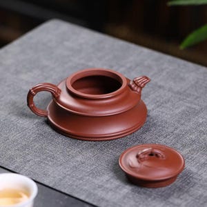 May include: A reddish-brown clay teapot with a wide, low-profile body, a bamboo-textured handle, and a matching lid placed beside it. The teapot features a short, curved spout and a smooth, matte finish, showcasing a traditional design.