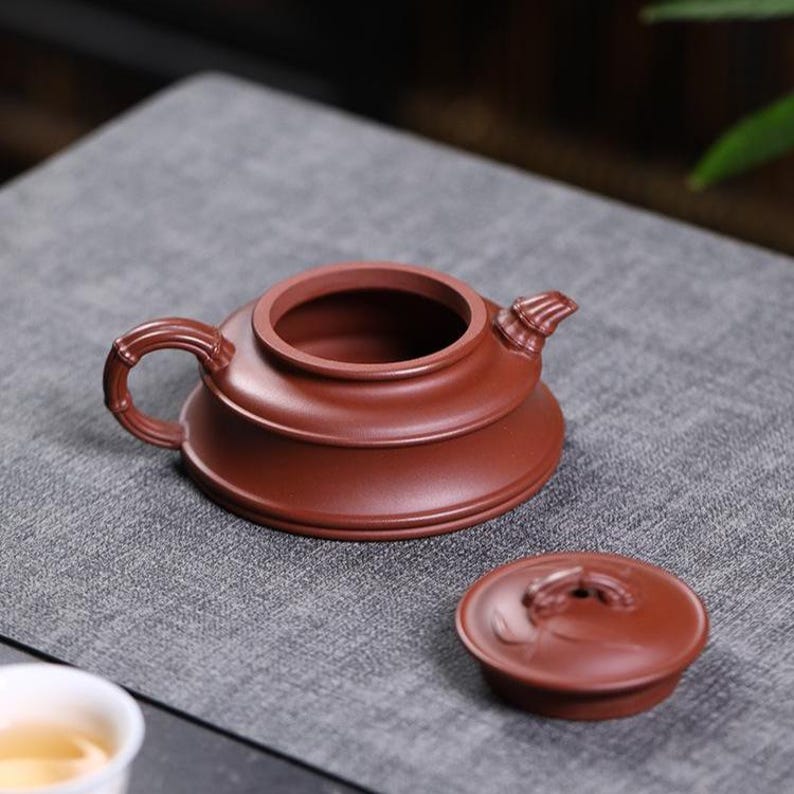 May include: A reddish-brown clay teapot with a wide, low-profile body, a bamboo-textured handle, and a matching lid placed beside it. The teapot features a short, curved spout and a smooth, matte finish, showcasing a traditional design.