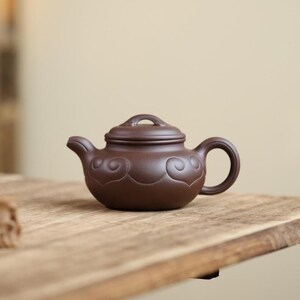 Op de afbeelding: Kleine, bruine keramische theepot met bijpassende deksel. De theepot heeft een afgeronde body, een gebogen handvat en een tuit. Decoratieve wolkenachtige patronen zijn in de body geëtst. De theepot staat op een houten oppervlak.