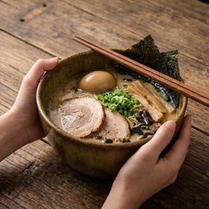 May include: A bowl of ramen with pork belly slices, a soft-boiled egg, green onions, and seaweed. The ramen is served in a brown ceramic bowl with wooden chopsticks resting on top. The bowl is held by two hands, set against a wooden table.