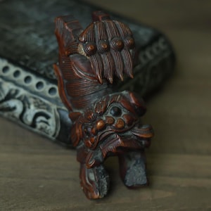 May include: A detailed, hand-carved wooden sculpture of a mythical creature. The sculpture is primarily brown with intricate details, including a clawed paw and a stylized face. The piece is set against a blurred background.