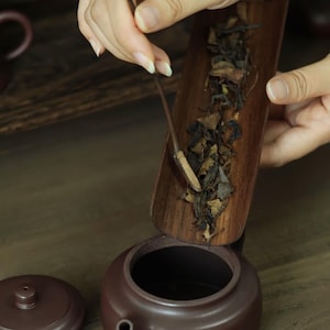 May include: A dark brown, matte-finished clay teapot with a matching lid placed beside it. A long, narrow, dark wooden tea scoop filled with dried, dark brown and tan tea leaves is being tilted to pour the leaves into the open teapot.