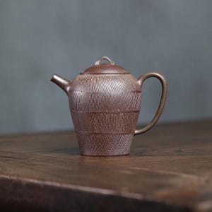Peut inclure: Une théière en céramique marron avec un motif texturé et nervuré. La théière a une anse incurvée, un bec verseur et un couvercle avec une petite boucle. La théière est posée sur une surface en bois.