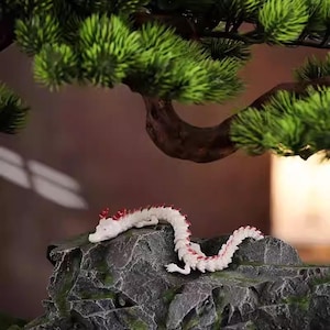 May include: A white and red dragon figurine rests on a gray, textured rock. The dragon has a detailed design with red accents along its spine. A green bonsai tree is in the background, adding a natural element to the scene.