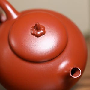 May include: A close-up of a reddish-brown ceramic teapot. The teapot has a rounded body, a small spout, and a lid with a decorative knob. The teapot is likely used for brewing tea.