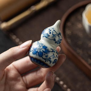 May include: A small, gourd-shaped ceramic vessel featuring a white base with intricate blue floral patterns and scrolling vine motifs. The base of the object is decorated with a dark blue and gold band, creating a distinct contrast against the white.