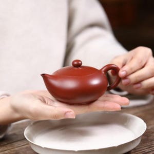 May include: A small, smooth, reddish-brown clay teapot with a rounded body, a short spout, a curved handle, and a matching lid with a small knob. The teapot has a matte finish and a simple, functional design for brewing tea.