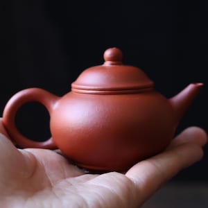 Könnte beinhalten: Kleine, rotbraune Keramik-Teekanne mit gebogenem Henkel und Deckel mit kleinem Knauf. Die Teekanne wird in einer offenen Hand vor dunklem Hintergrund gehalten. Das Design ist schlicht und klassisch, passend für Teezeremonien.