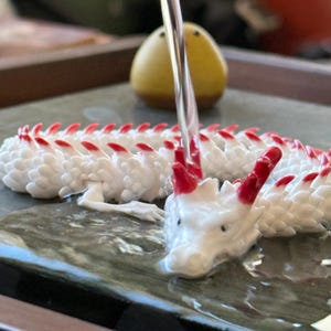 May include: A white and red ceramic dragon figurine with water pouring over it. The dragon has detailed scales and red accents on its horns and spine. A yellow, pear-shaped object is in the background.
