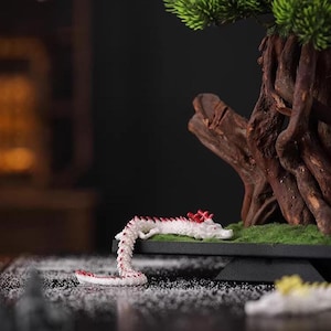 May include: A miniature white dragon figurine with red accents, resting on a small, black platform with green moss. The dragon is positioned near a bonsai tree with a dark background.