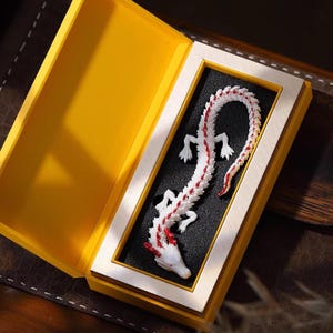May include: A white and red dragon figurine inside a yellow box with a black interior. The dragon has a detailed design with red accents on its head, tail, and spine. The box is open, revealing the dragon's full length.