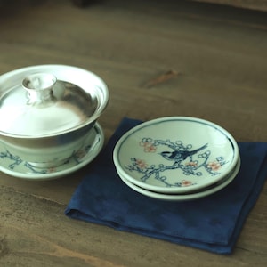 May include: A silver-coloured lidded tea bowl with a matching saucer featuring blue and orange floral patterns, alongside two small white ceramic saucers decorated with a blue bird perched on a flowering branch with orange blossoms.