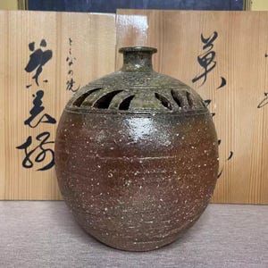 Puede incluir: Un jarrón de cerámica artesanal y moteado con un esmalte marrón oscuro y verde. El jarrón tiene un cuerpo redondeado, un cuello estrecho y una parte superior decorativa perforada. El fondo incluye paneles de madera con caligrafía japonesa.