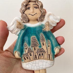 May include: A ceramic angel figurine with a teal and white crackle glaze finish. The angel features textured brown hair, white wings, and a central graphic depicting a cluster of buildings with pointed roofs. The base is white with crackle details.