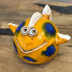 May include: A round ceramic sculpture of a fish with a bright yellow body, dark blue circular spots, and white textured fins. The fish features two prominent, wide-set eyes with dark pupils and a carved horizontal line representing a smiling mouth.