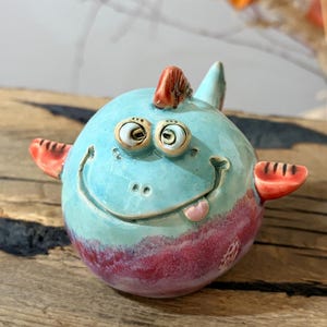 Può includere: Una statuetta rotonda in ceramica a forma di creatura con la parte superiore azzurra e quella inferiore rosa scuro. Presenta due grandi occhi sporgenti, un'ampia bocca sorridente con una piccola lingua che sporge e pinne arancioni sui lati.