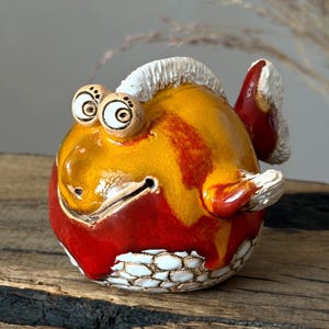 Könnte beinhalten: Eine runde Keramik-Fischfigur mit einem glänzend gelben Oberkörper und einem tiefroten Unterteil. Der Fisch hat große, hervorstehende Augen mit schwarzen Pupillen, eine strukturierte weiße Rückenflosse, eine weiße Schwanzflosse und weiße Schuppen.