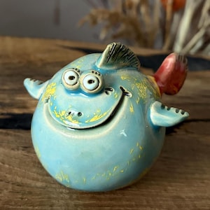 May include: A round ceramic fish figurine featuring a light blue glaze with yellow accents. The fish has two prominent protruding eyes, a wide smiling mouth, a dorsal fin on top, two side fins, and a distinct red tail fin extending from the back.
