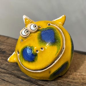May include: A whimsical ceramic sculpture of a round, yellow creature with large, googly eyes and small horns. The creature has blue and green spots and a smiling mouth. The sculpture is placed on a wooden surface.
