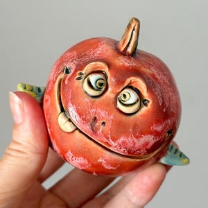 Puede incluir: Una escultura de cerámica roja y redonda que presenta un rostro expresivo con ojos grandes, una amplia sonrisa y una pequeña protuberancia en la parte superior. La superficie tiene un esmalte brillante y moteado con reflejos blancos y aletas azules.