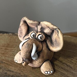 May include: A small, hand-sculpted clay elephant figurine with a textured, earthy brown finish. The figure features large, floppy ears, two prominent white tusks, and wide, expressive eyes with dark pupils. It sits in a rounded, compact pose.