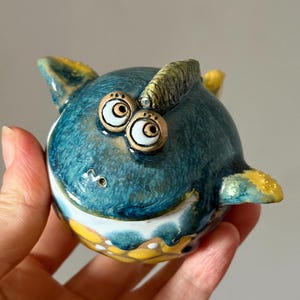 Op de afbeelding: Een rond keramieken beeldje in de vorm van een wezen met een donkerblauw getextureerd lichaam, twee prominente uitpuilende ogen en gele accenten op de vinnen en de basis. Het oppervlak heeft een glanzende afwerking met zichtbare penseelstreken.