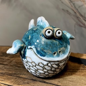 May include: A whimsical ceramic fish sculpture with a round body and large, expressive eyes. The fish features a blue and white glazed finish with a textured pattern on the belly. It has small fins and a smiling mouth, creating a playful aesthetic.