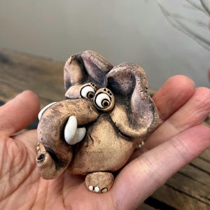 May include: A whimsical ceramic elephant figurine with large, expressive eyes and oversized ears. The elephant is a light brown color with white tusks and small feet. The figurine is held in a hand.