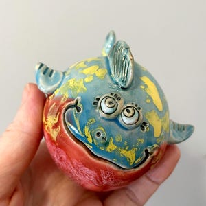 Puede incluir: Una figura de pez de cerámica redonda con un acabado brillante y una paleta de colores vibrantes en azul cielo, amarillo brillante y rojo intenso. La criatura tiene dos ojos grandes y separados, una boca curva y aletas pequeñas.